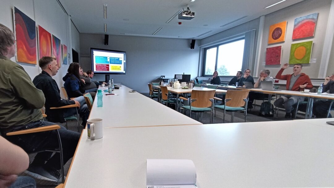 Das Foto zeigt mehrere Personen in eine Konferenzraum. Sie diskutieren augenscheinlich. Einige schauen zum Webex-Board, auf dem eine Präsentation gezeigt wird.