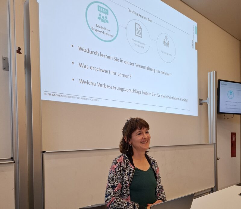 Eine Dozentin steht lächelnd vor einer Präsentationsfolie in einem Seminarraum der FH Aachen. Auf der Folie ist das Verfahren des "Teaching Analysis Poll" (TAP) dargestellt, das aus einer moderierten Gruppendiskussion, einem anonymisierten TAP-Bericht und einem Nachgespräch besteht. Darunter stehen drei Leitfragen zur Verbesserung der Lehrveranstaltung: Wodurch lernen Sie in dieser Veranstaltung am meisten? Was erschwert Ihr Lernen? Welche Verbesserungsvorschläge haben Sie für die hinderlichen Punkte? Rechts im Bild ist ein zweiter Bildschirm mit identischer Darstellung sichtbar. Die Präsentation ist mit dem Logo der FH Aachen – University of Applied Sciences – versehen.