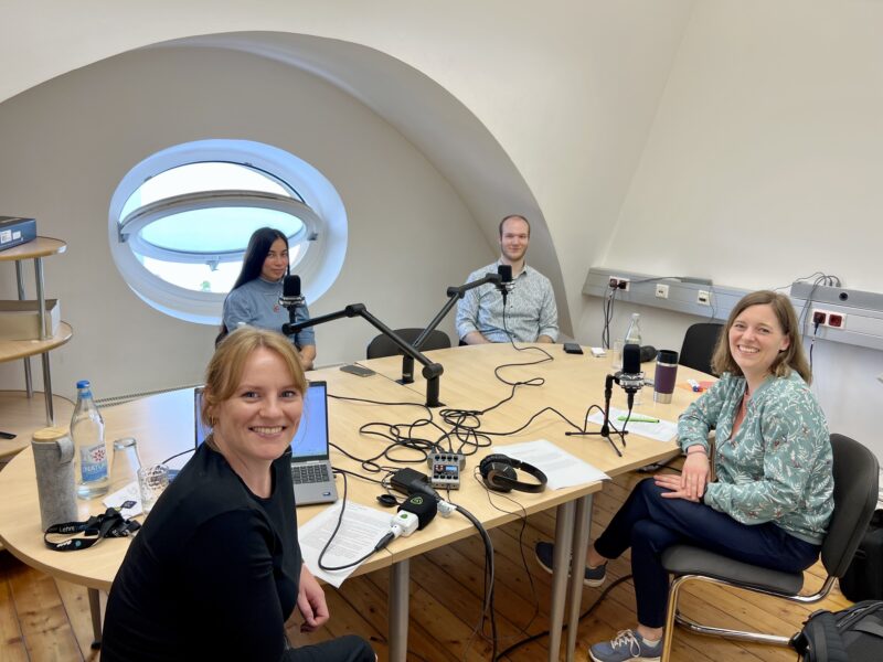 Vier Personen sitzen an einem runden Tisch mit Mikrofonen und Aufnahmegeräten und nehmen gemeinsam eine Podcastfolge auf. Zwei Frauen sitzen vorne im Bild und lächeln in die Kamera, im Hintergrund sitzen eine Frau und ein Mann vor Mikrofonen.