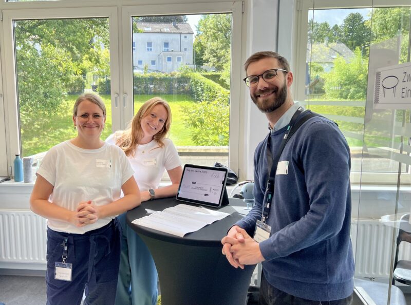 Drei Personen stehen freundlich lächelnd an einem Stehtisch in einem hellen Raum. Auf dem Tisch liegen Unterlagen und ein Tablet mit der Aufschrift „Tag der Lehre 2025“. Hinter ihnen sind große Fenster mit Blick auf eine grüne Wiese und Gebäude zu sehen.